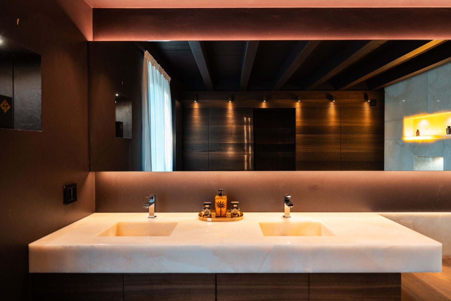 Modern bathroom interior featuring a sleek double sink with a marble countertop, stylish faucets, and ambient lighting. The dark walls contrast with wooden accents, while natural light filters through sheer curtains. Decorative elements include a stylish tray with toiletries. | Only on SopranoVillas