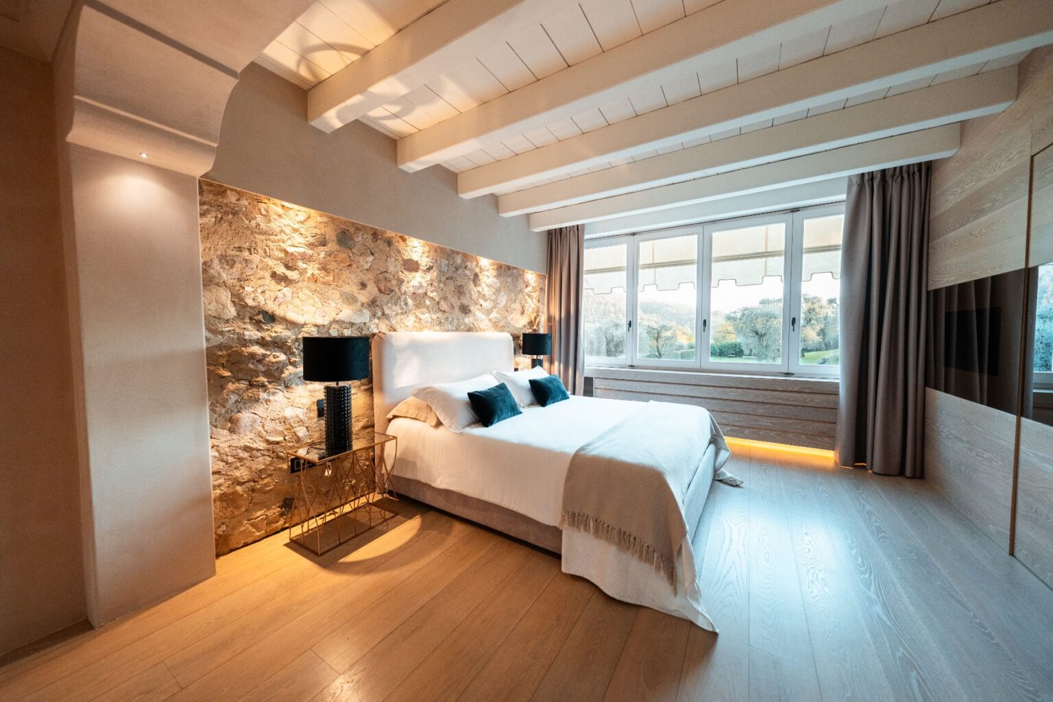 Modern bedroom with a stone accent wall, featuring a king-sized bed dressed in white linens and dark blue pillows. The room includes stylish black bedside lamps, large windows with curtains, and warm wooden flooring, creating a cozy and inviting atmosphere. | Only on SopranoVillas