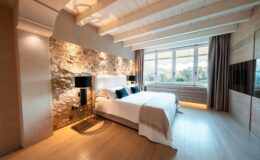 Modern bedroom with a stone accent wall, featuring a king-sized bed dressed in white linens and dark blue pillows. The room includes stylish black bedside lamps, large windows with curtains, and warm wooden flooring, creating a cozy and inviting atmosphere.