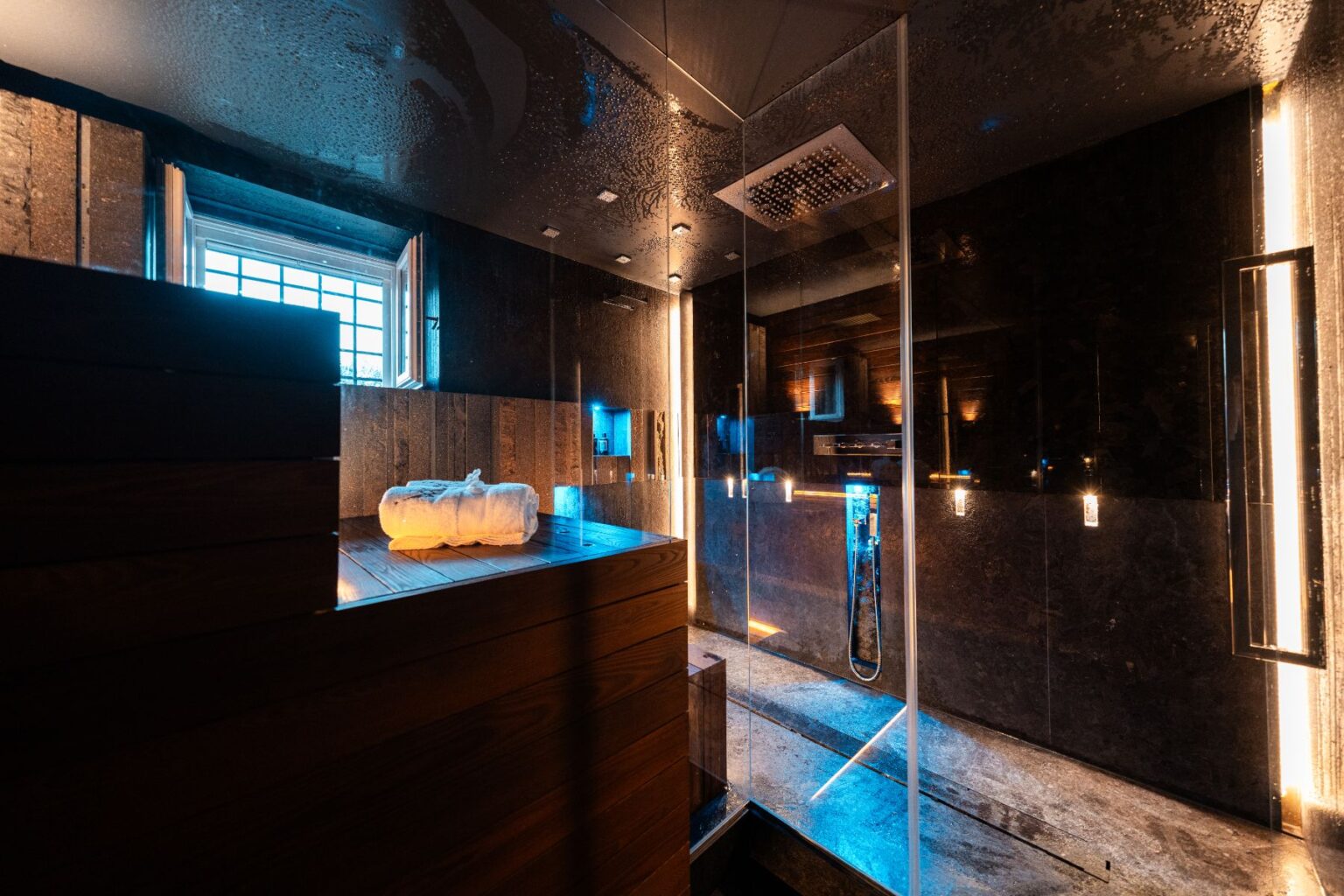 Modern spa bathroom featuring a glass shower, wooden sauna, and ambient lighting. Soft towels are neatly arranged on a wooden bench, creating a serene and luxurious atmosphere for relaxation. | Only on SopranoVillas