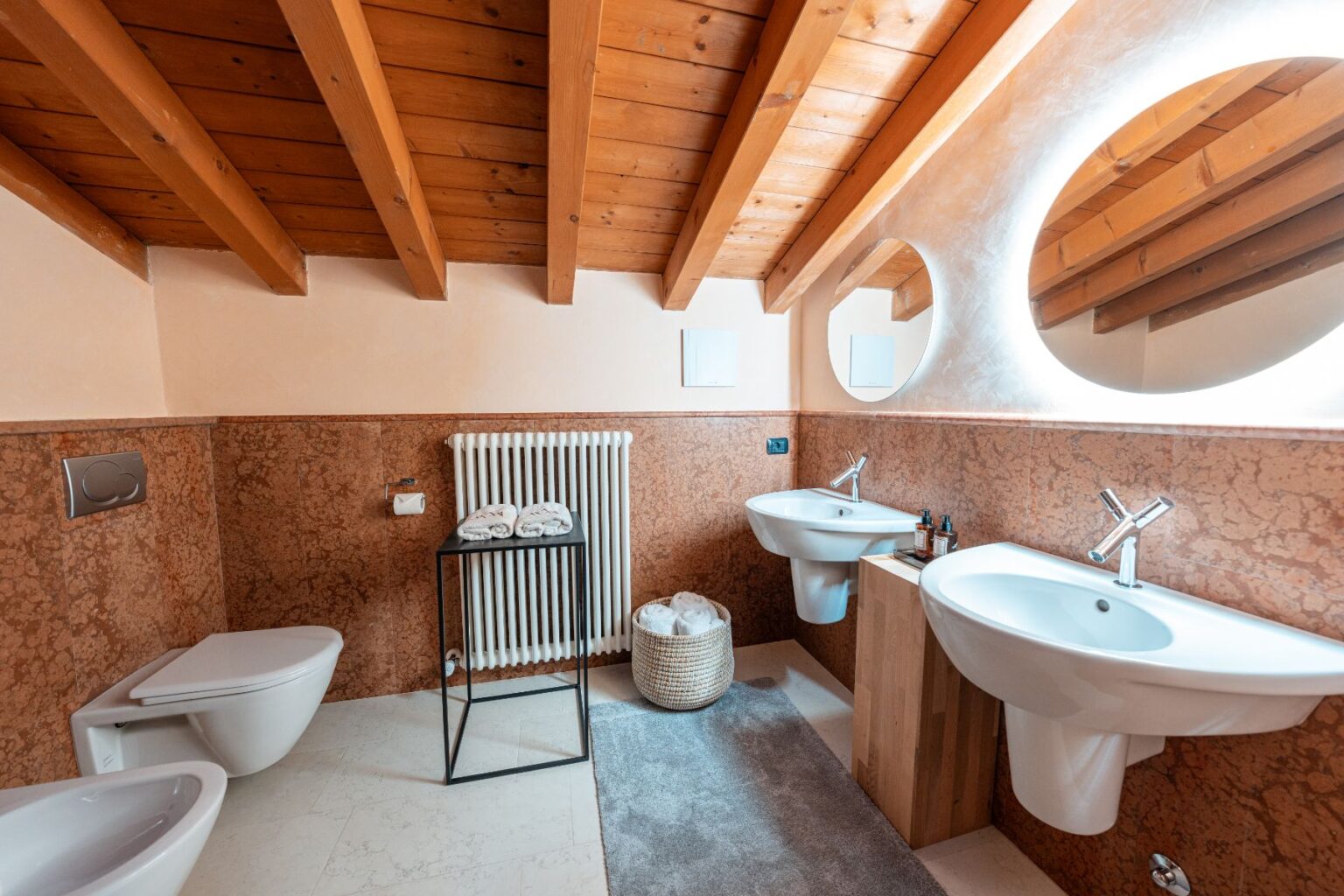 Modern bathroom interior featuring wooden beams, dual sinks, a stylish mirror, and a cozy atmosphere. The design includes a heated towel rack, decorative towels, and a woven basket, creating a functional and aesthetic space. | Only on SopranoVillas