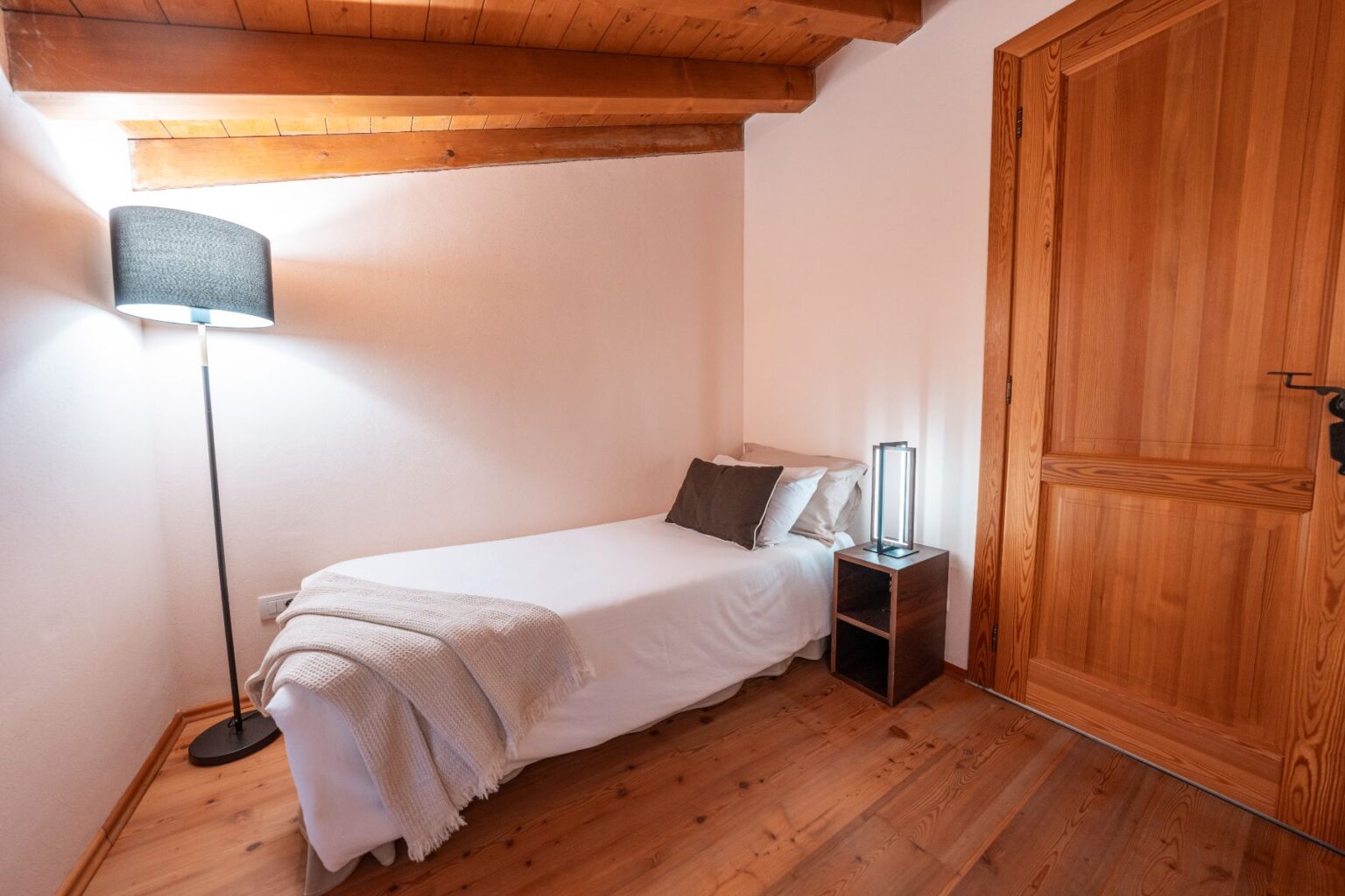 Cozy bedroom featuring a single bed with a white blanket, accented by decorative pillows. A stylish floor lamp and a modern bedside table enhance the inviting atmosphere, complemented by warm wooden beams and flooring. | Only on SopranoVillas