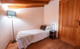 Cozy bedroom featuring a single bed with a white blanket, accented by decorative pillows. A stylish floor lamp and a modern bedside table enhance the inviting atmosphere, complemented by warm wooden beams and flooring.