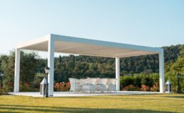 Modern outdoor pergola with white furniture set, surrounded by lush greenery and colorful flowers, ideal for relaxation and gatherings.