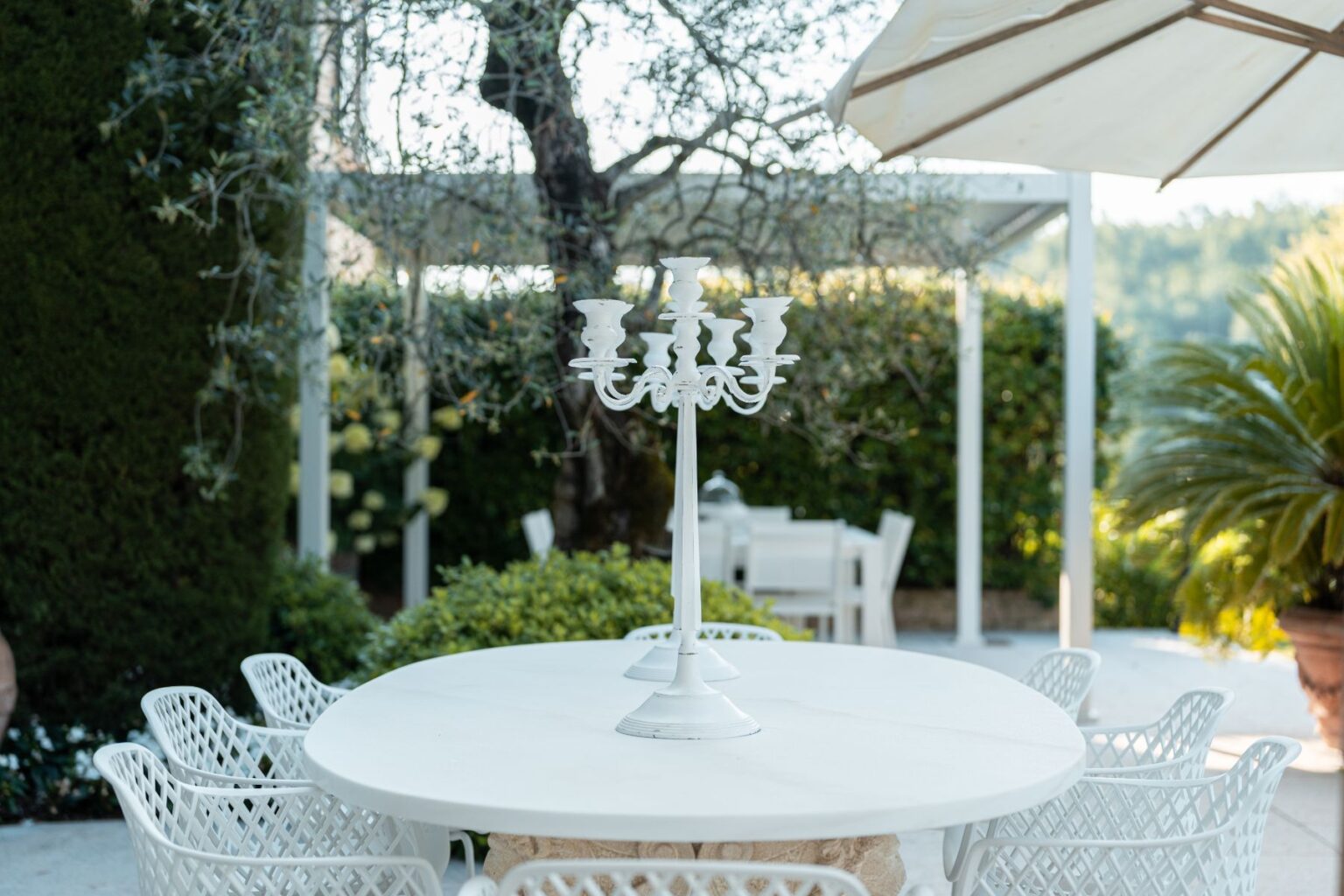 Outdoor dining area featuring a round white table with a decorative white candelabra at the center, surrounded by stylish white chairs, set against a lush green garden backdrop. | Only on SopranoVillas