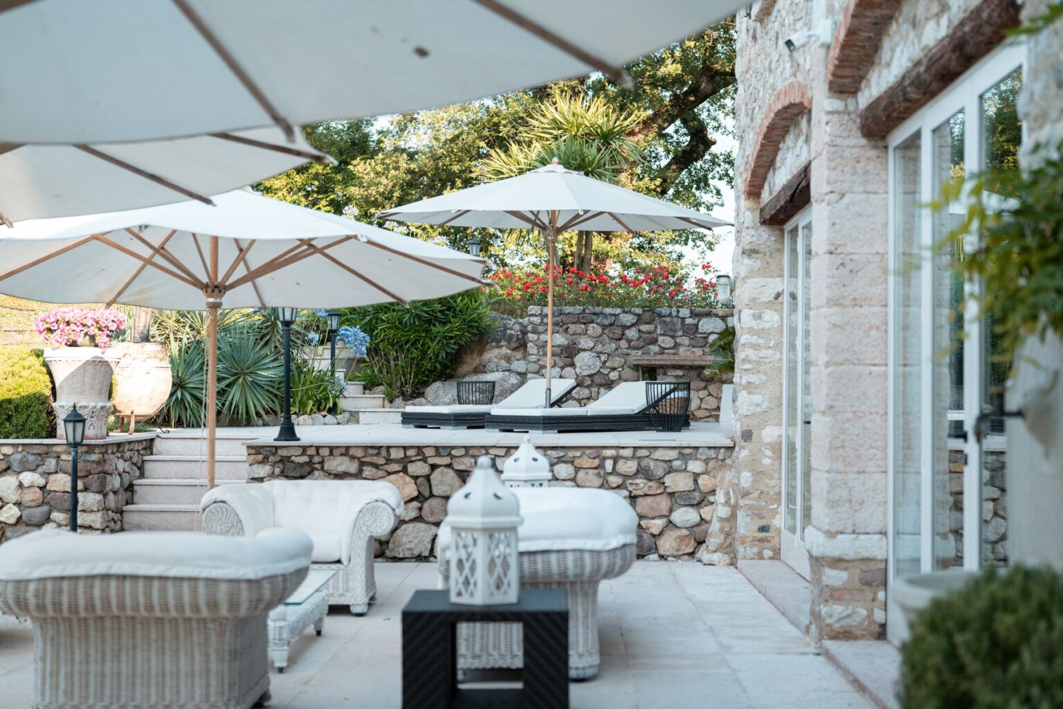 Outdoor patio area featuring white sun loungers under large umbrellas, surrounded by lush greenery and colorful flowers. The space includes stylish wicker furniture and stone walls, creating a serene and inviting atmosphere for relaxation. | Only on SopranoVillas