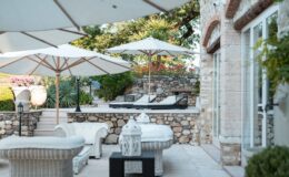 Outdoor patio area featuring white sun loungers under large umbrellas, surrounded by lush greenery and colorful flowers. The space includes stylish wicker furniture and stone walls, creating a serene and inviting atmosphere for relaxation.