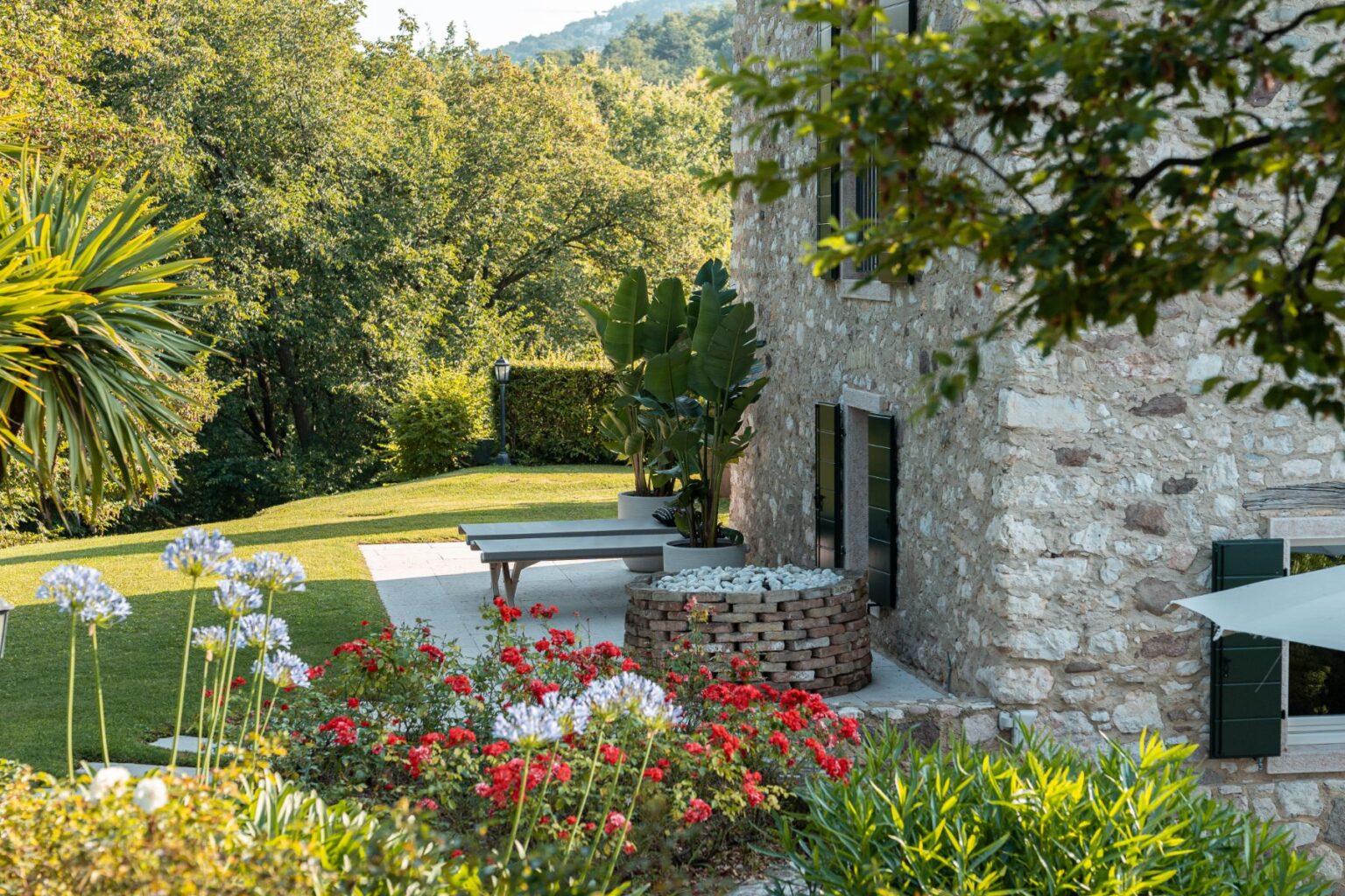Charming stone house surrounded by lush greenery and vibrant flower beds, featuring a modern outdoor seating area and decorative stone planter in a serene garden setting. | Only on SopranoVillas