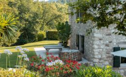 Charming stone house surrounded by lush greenery and vibrant flower beds, featuring a modern outdoor seating area and decorative stone planter in a serene garden setting.