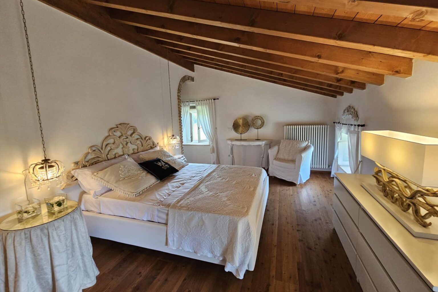 Cozy bedroom featuring a king-sized bed with an ornate headboard, decorative pillows, and stylish pendant lights. The room has wooden beams, warm wooden flooring, and elegant furnishings including a white armchair and a decorative side table. Natural light filters through the window, enhancing the inviting atmosphere. | Only on SopranoVillas