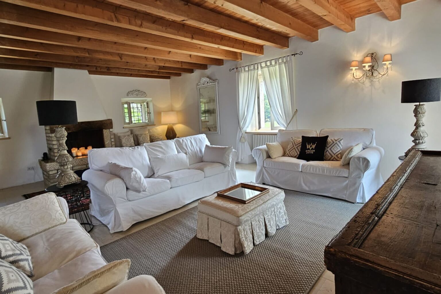 Cozy living room featuring white slipcovered sofas, a decorative coffee table, and wooden beams on the ceiling. The space is illuminated by natural light from large windows and soft lamps, creating a warm and inviting atmosphere. A fireplace with candles adds a touch of charm to the decor. | Only on SopranoVillas