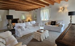 Cozy living room featuring white slipcovered sofas, a decorative coffee table, and wooden beams on the ceiling. The space is illuminated by natural light from large windows and soft lamps, creating a warm and inviting atmosphere. A fireplace with candles adds a touch of charm to the decor.