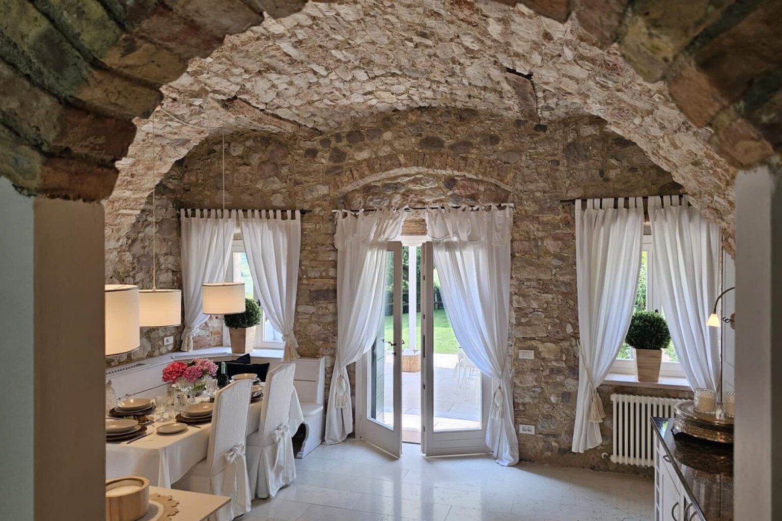 Cozy kitchen interior featuring stone walls and an arched ceiling, adorned with white curtains and modern pendant lights. A dining table is set with elegant dishware, flowers, and a bottle of wine, while double doors open to a green outdoor space. Perfect for a rustic yet stylish home ambiance. | Only on SopranoVillas