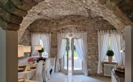 Cozy kitchen interior featuring stone walls and an arched ceiling, adorned with white curtains and modern pendant lights. A dining table is set with elegant dishware, flowers, and a bottle of wine, while double doors open to a green outdoor space. Perfect for a rustic yet stylish home ambiance.