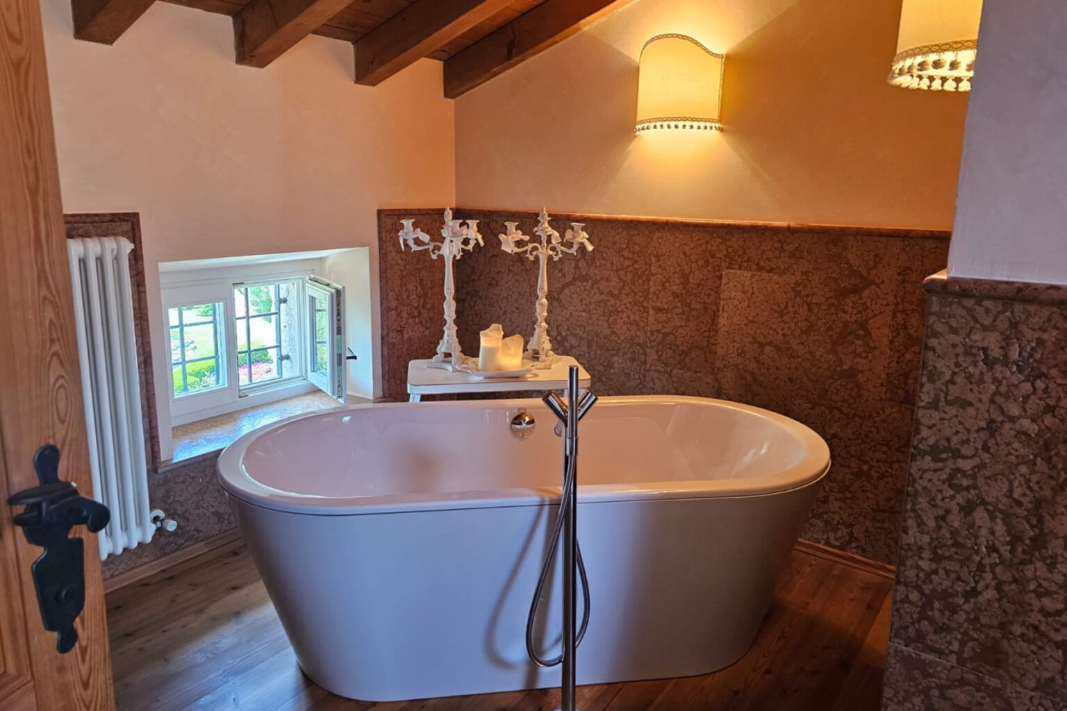 Modern bathroom interior featuring a freestanding white bathtub, elegant lighting, and a scenic window view. The warm wooden beams and decorative wall add a cozy touch, complemented by a stylish candle holder on a small table. | Only on SopranoVillas