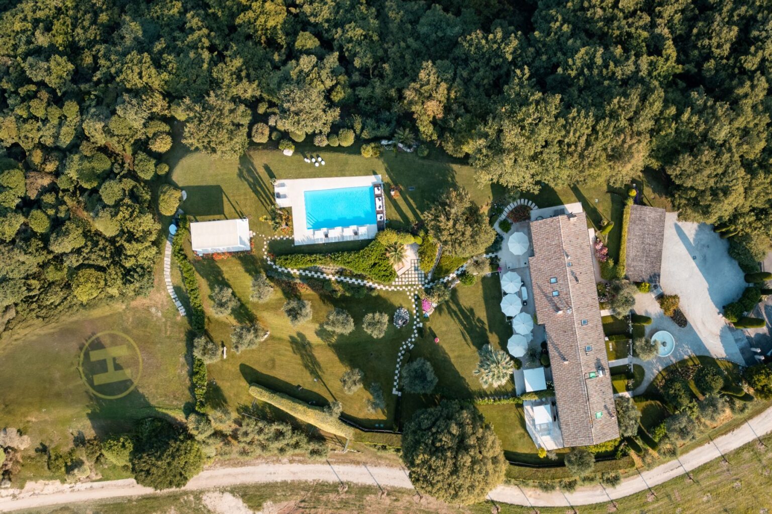 Aerial view of a luxurious villa surrounded by lush greenery, featuring a sparkling blue swimming pool, landscaped gardens, and a helipad. The property includes a spacious main house with a tiled roof and outdoor seating areas, ideal for relaxation and entertainment. | Only on SopranoVillas