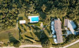 Aerial view of a luxurious villa surrounded by lush greenery, featuring a sparkling blue swimming pool, landscaped gardens, and a helipad. The property includes a spacious main house with a tiled roof and outdoor seating areas, ideal for relaxation and entertainment.