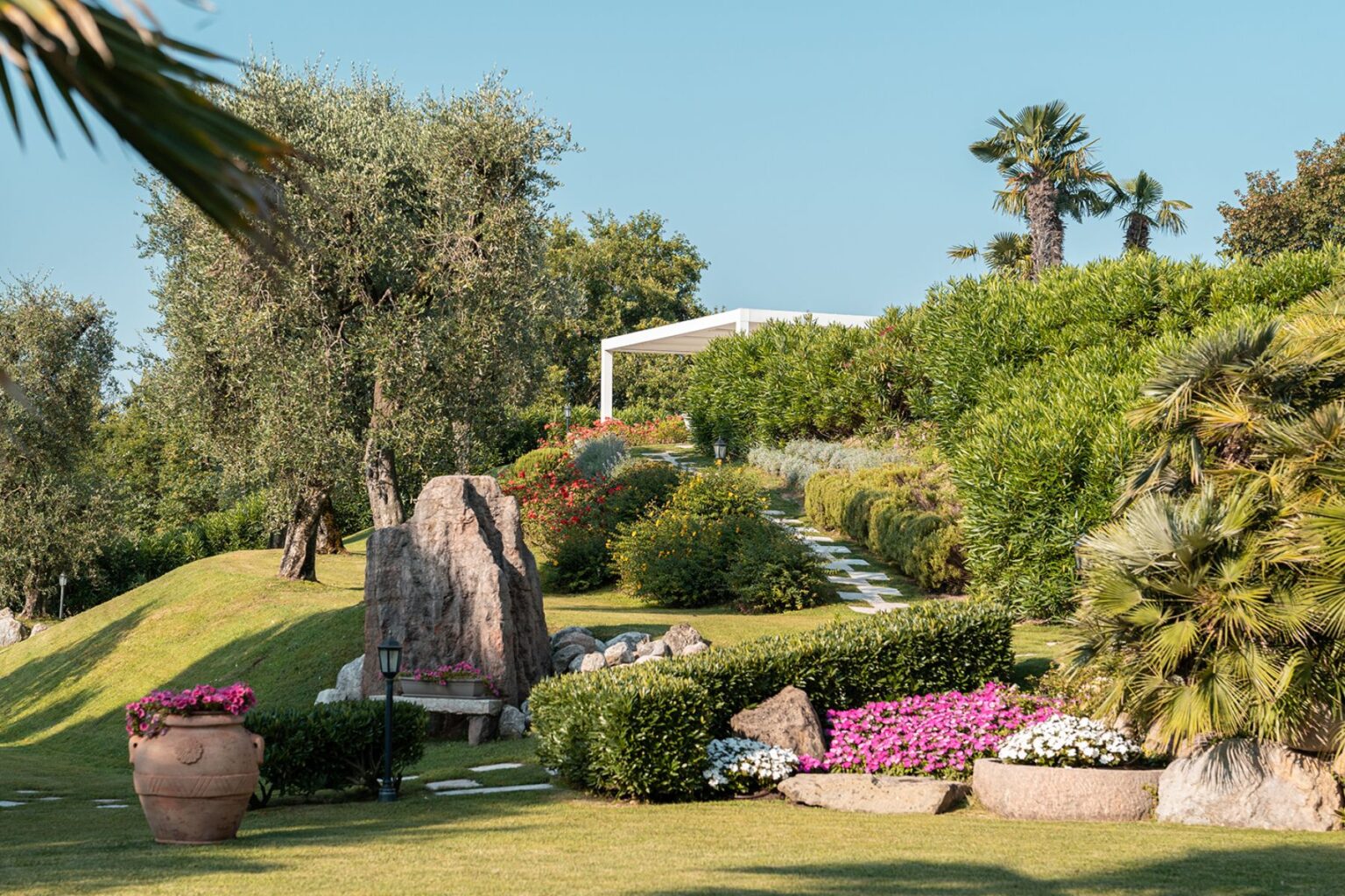 Lush garden landscape featuring vibrant flowers, olive trees, and a stone pathway leading to a modern outdoor pavilion, surrounded by greenery and decorative rocks. Ideal for outdoor relaxation and landscaping inspiration. | Only on SopranoVillas