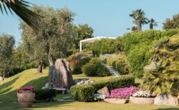 Lush garden landscape featuring vibrant flowers, olive trees, and a stone pathway leading to a modern outdoor pavilion, surrounded by greenery and decorative rocks. Ideal for outdoor relaxation and landscaping inspiration.
