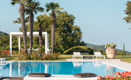 Luxurious outdoor swimming pool surrounded by vibrant greenery and palm trees, featuring white sun loungers and a decorative planter, under a clear blue sky. Ideal for relaxation and leisure in a tranquil garden setting.
