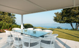 Outdoor dining area featuring a glass table surrounded by white wicker chairs, set against a scenic view of lush greenery and a distant body of water. Ideal for entertaining or relaxing in a serene environment.