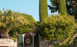 Charming garden entrance featuring stone walls, green wooden doors, and lush landscaping with palm trees and tall cypress trees under a clear blue sky.