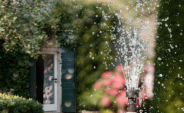 Charming garden scene featuring a decorative fountain spraying water, with a stone house and green shutters in the background, surrounded by lush greenery and colorful flowers.