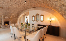 Elegant dining area with a glass table surrounded by beige upholstered chairs, set under a vaulted stone ceiling. Features include a decorative fireplace, ornate mirrors, and a stylish chandelier, enhancing the sophisticated ambiance.