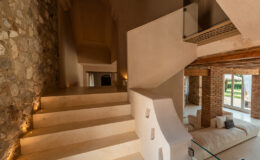 Spacious interior staircase leading to a well-lit living area, featuring a blend of natural stone and smooth plaster walls, with modern glass railings and cozy seating.