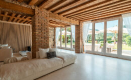 Spacious modern living room featuring exposed wooden beams and brick walls, with large glass doors leading to a lush garden area. The room includes a cozy white sofa, decorative pillows, and a stylish coffee table with a magazine.