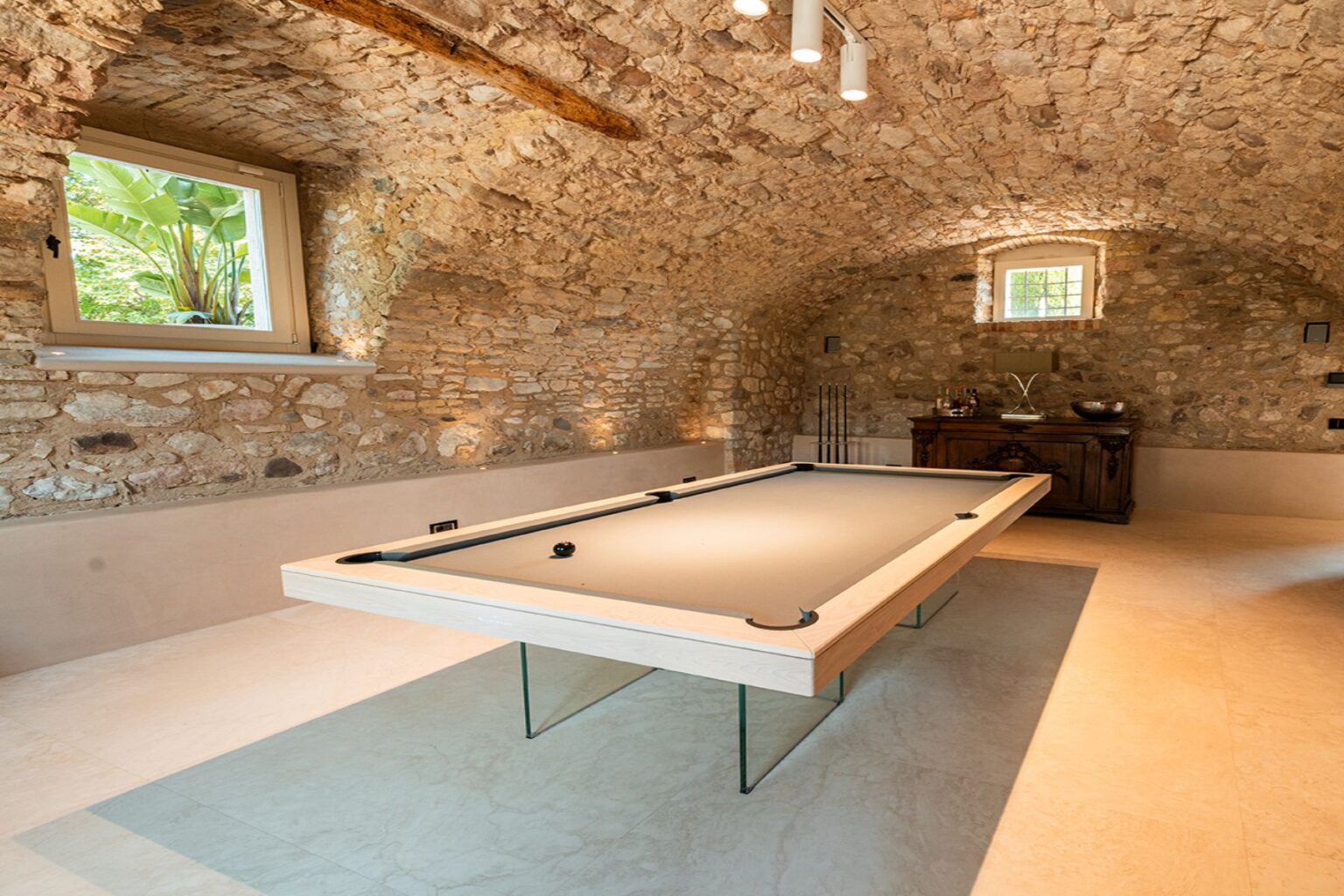 Elegant game room featuring a modern pool table with a sleek design, set against rustic stone walls and an arched ceiling. Natural light streams through a window surrounded by greenery, enhancing the inviting atmosphere. A stylish wooden cabinet is visible in the background, adding to the room's charm. | Only on SopranoVillas
