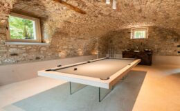 Elegant game room featuring a modern pool table with a sleek design, set against rustic stone walls and an arched ceiling. Natural light streams through a window surrounded by greenery, enhancing the inviting atmosphere. A stylish wooden cabinet is visible in the background, adding to the room's charm.