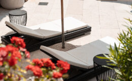 Luxurious outdoor patio featuring two white lounge chairs with an umbrella, surrounded by vibrant red flowers and modern black side tables, ideal for relaxation in a stylish setting.