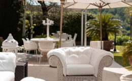 Elegant outdoor patio featuring white upholstered chairs, a glass table, and large umbrellas providing shade. Lush greenery and a dining area with stylish chairs create a serene atmosphere for relaxation and entertaining.