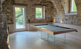 Modern game room featuring a sleek glass and wood pool table, surrounded by rustic stone walls and arched ceilings, with large windows providing natural light and views of the garden.