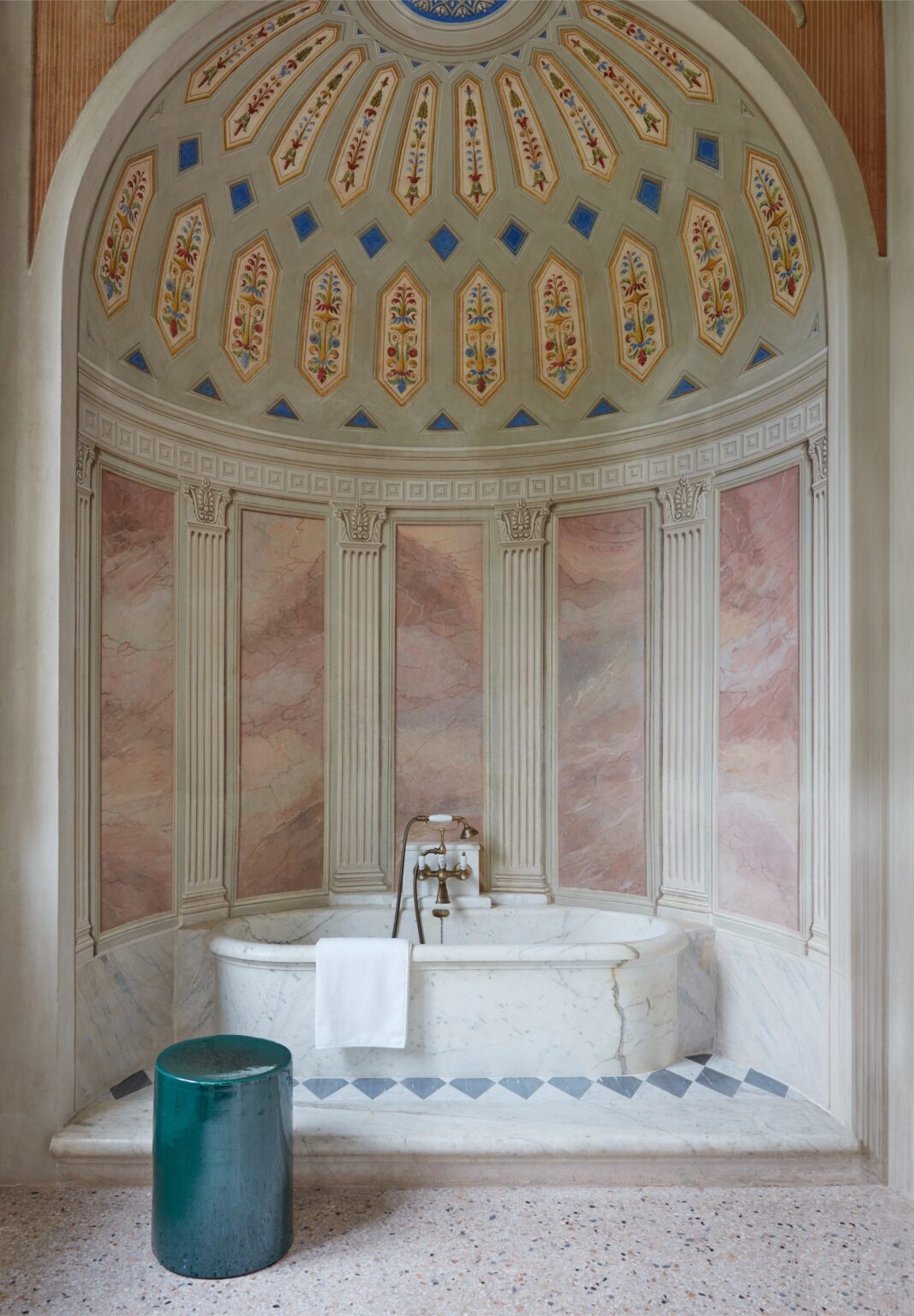 Elegant bathroom interior featuring a marble bathtub, decorative plaster walls with floral motifs, and a green stool. The space showcases classical design elements, including fluted columns and a beautifully painted ceiling, creating a serene and luxurious atmosphere. | Only on SopranoVillas