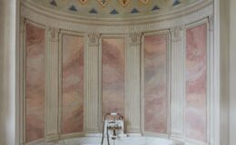 Elegant bathroom interior featuring a marble bathtub, decorative plaster walls with floral motifs, and a green stool. The space showcases classical design elements, including fluted columns and a beautifully painted ceiling, creating a serene and luxurious atmosphere.