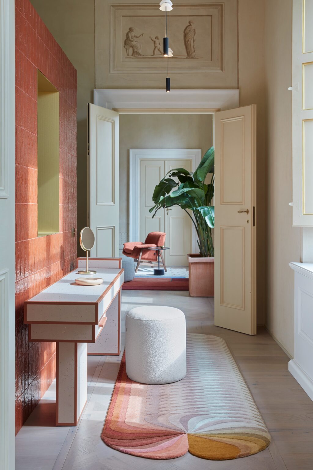 Modern hallway interior featuring a stylish console table with a round mirror, a plush white ottoman, and a colorful area rug. The space is illuminated by natural light, showcasing a vibrant red accent wall and lush green plants, with elegant doors leading to additional rooms. | Only on SopranoVillas