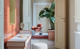 Modern hallway interior featuring a stylish console table with a round mirror, a plush white ottoman, and a colorful area rug. The space is illuminated by natural light, showcasing a vibrant red accent wall and lush green plants, with elegant doors leading to additional rooms.