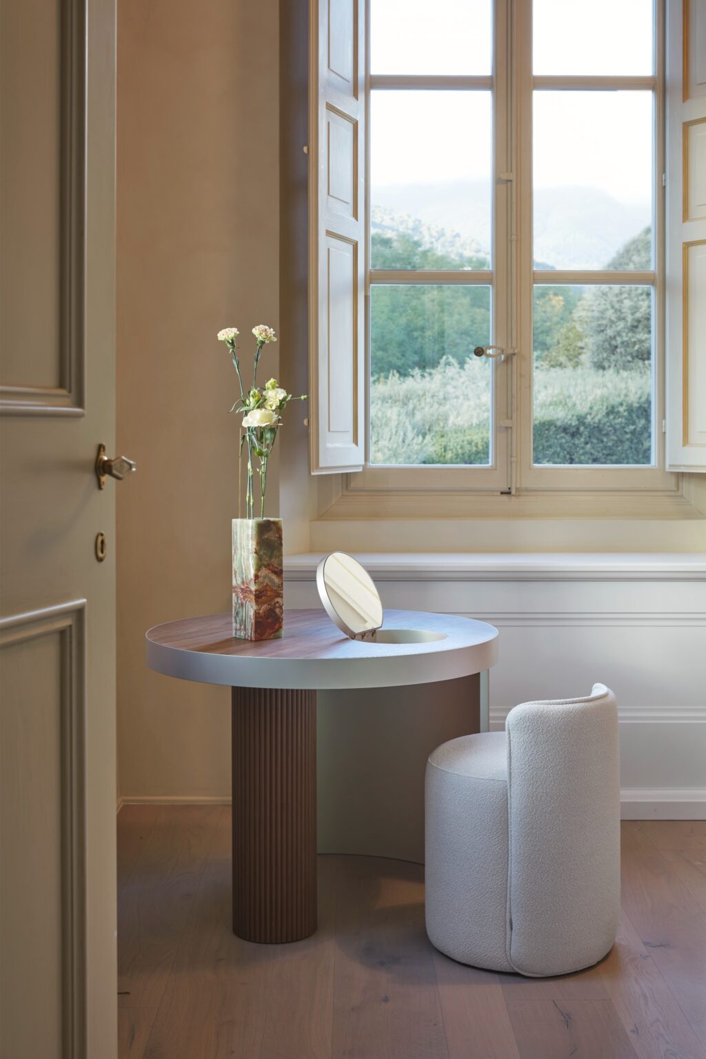 Modern interior design featuring a round table with a sleek metal edge, a decorative vase with flowers, and a contemporary upholstered stool. The scene is illuminated by natural light streaming through a large window, showcasing a serene outdoor view. | Only on SopranoVillas