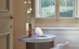 Modern interior design featuring a round table with a sleek metal edge, a decorative vase with flowers, and a contemporary upholstered stool. The scene is illuminated by natural light streaming through a large window, showcasing a serene outdoor view.