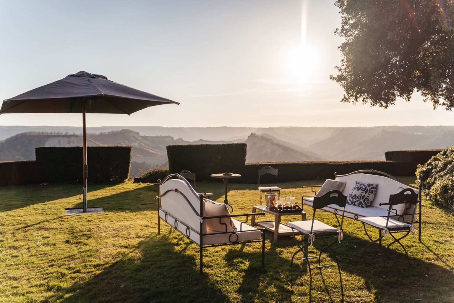 Elegant outdoor seating area with a stylish umbrella, set against a scenic mountain landscape during sunset. The arrangement includes a sofa, chairs, a small table, and a tray with glasses, creating a perfect spot for relaxation and enjoying nature. | Only on SopranoVillas