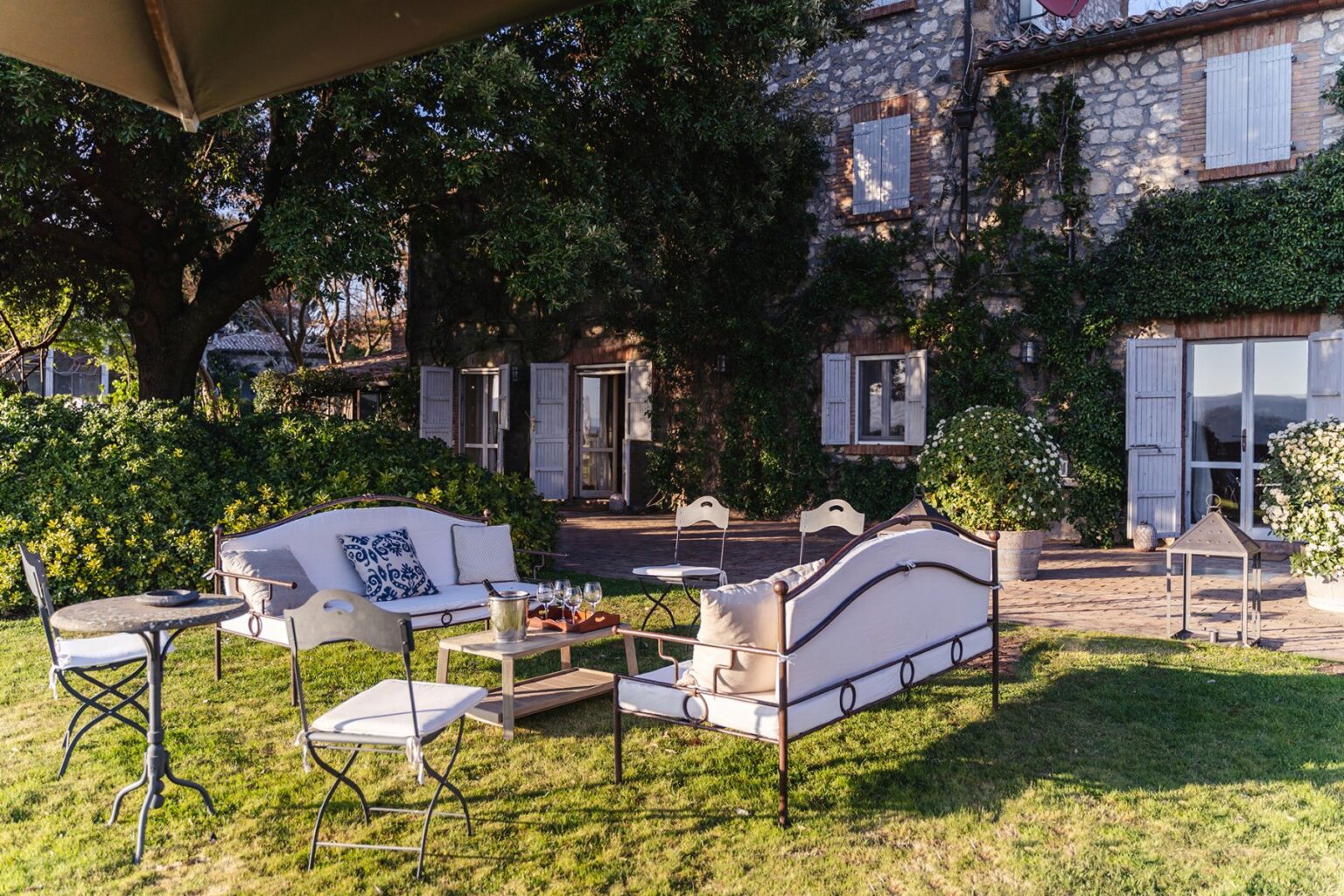Outdoor seating area with stylish furniture, including a white sofa and metal chairs, set against a charming stone house surrounded by greenery and blooming flowers. Ideal for relaxing or entertaining in a serene garden environment. | Only on SopranoVillas
