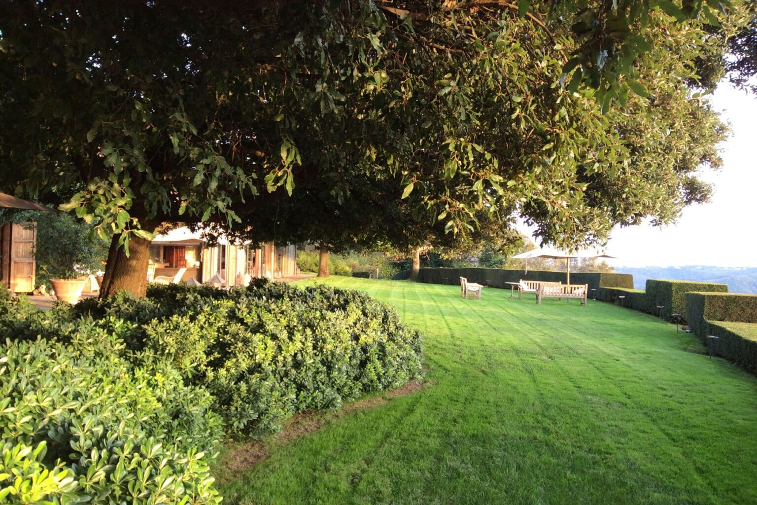 Lush green lawn with neatly trimmed hedges, shaded by a large tree, featuring outdoor seating and a modern home in the background, ideal for relaxation and outdoor gatherings. | Only on SopranoVillas