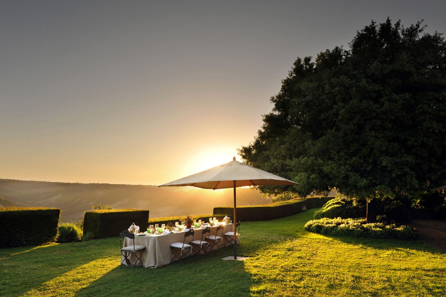 Outdoor dining setup under a large umbrella during sunset, featuring a beautifully arranged table with elegant glassware and tableware, surrounded by lush greenery and rolling hills in the background. | Only on SopranoVillas
