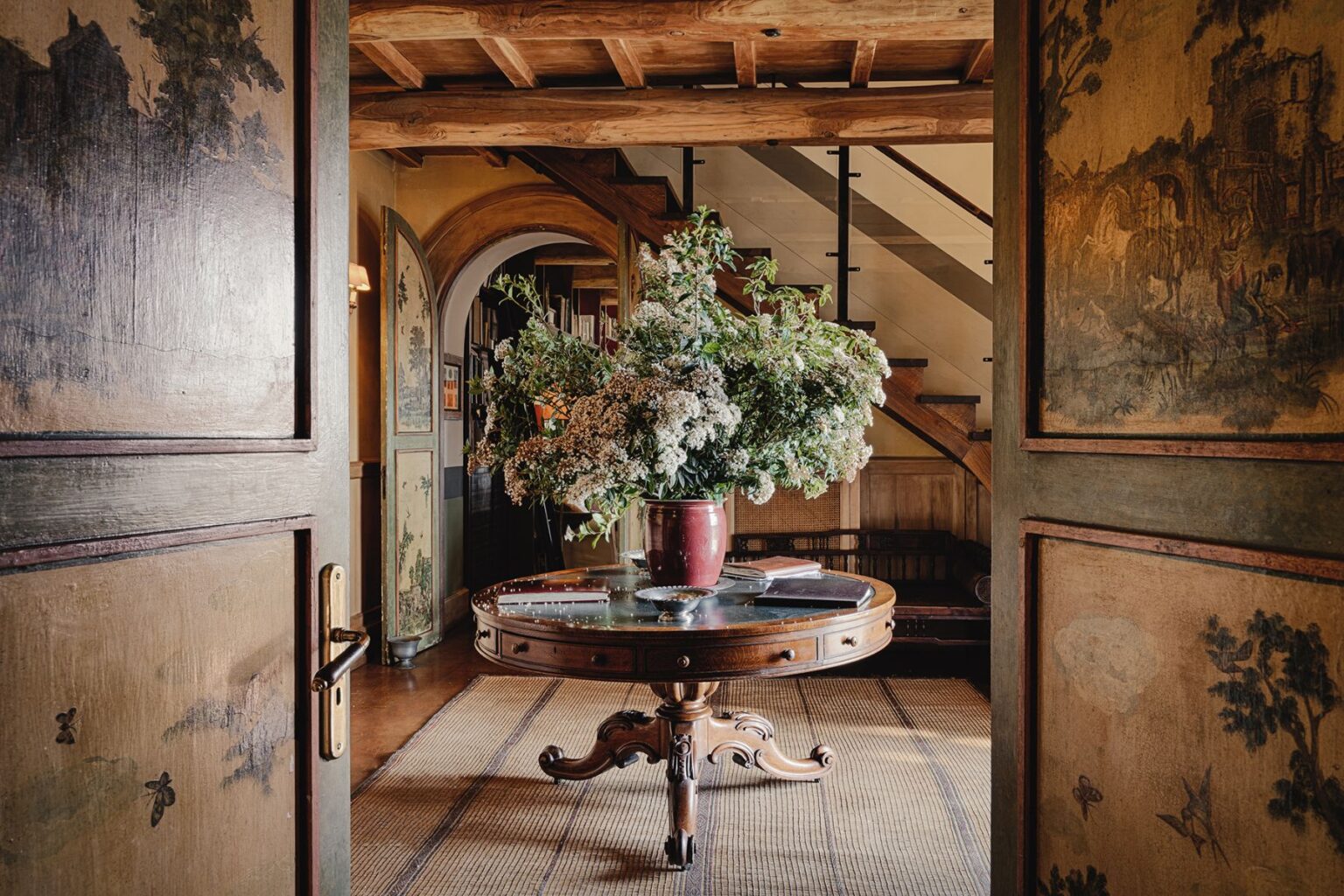 Interior view of a welcoming foyer featuring a round wooden table adorned with a large floral arrangement in a vase, surrounded by decorative walls and an elegant staircase in the background. The space showcases a blend of rustic charm and artistic details. | Only on SopranoVillas