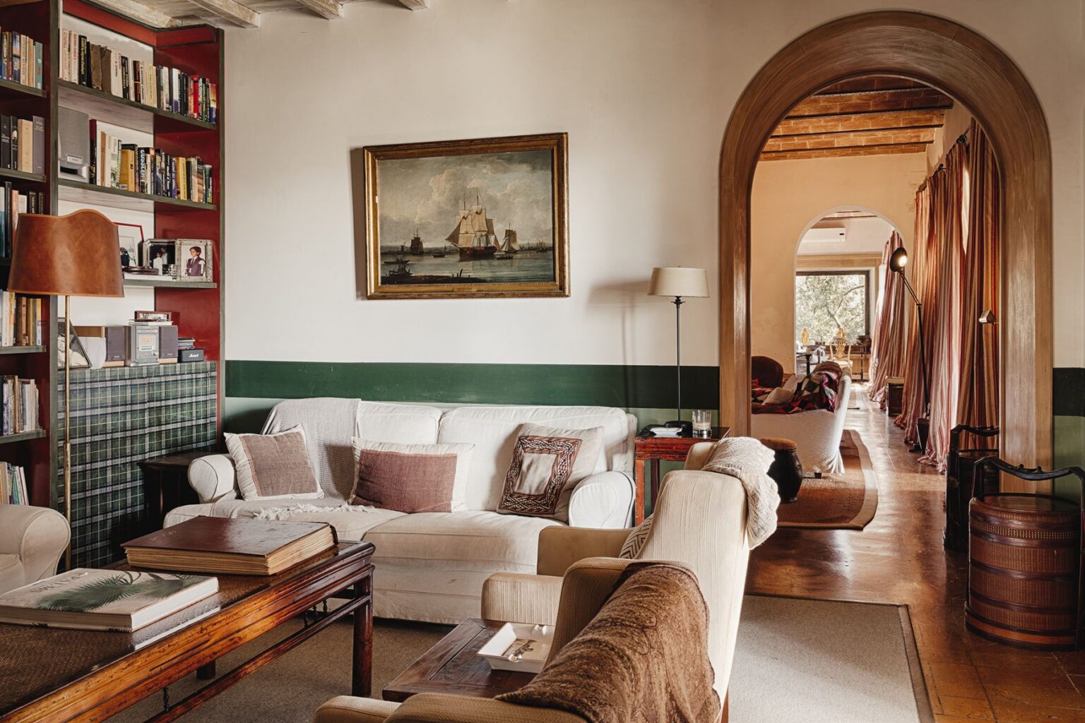 Cozy living room featuring a white sofa with decorative pillows, a wooden coffee table with books, and a bookshelf filled with books. The room includes a vintage painting of ships, a floor lamp, and an archway leading to another room with large windows and draped curtains. The interior design combines rustic and classic elements, creating a warm and inviting atmosphere. | Only on SopranoVillas