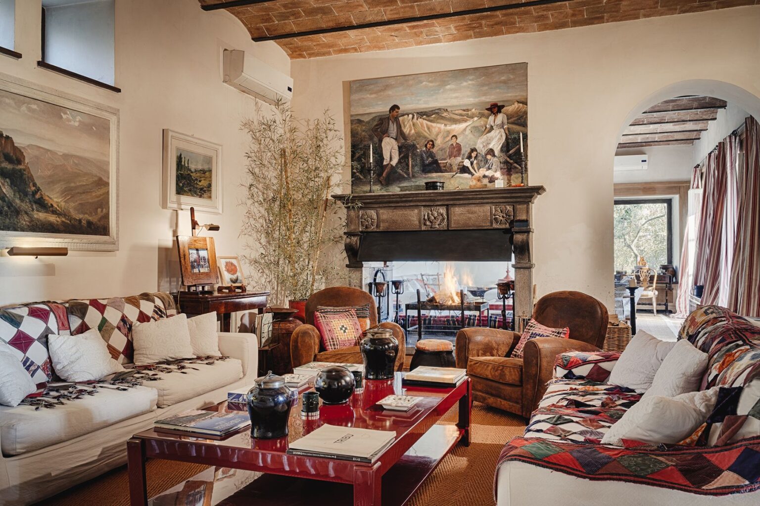 Cozy living room featuring a stone fireplace, vintage furniture, and artistic decor. The space includes two comfortable armchairs, a white sofa with a patchwork throw, and a red coffee table adorned with books and decorative items. Natural light filters through large windows, highlighting the warm, inviting atmosphere. | Only on SopranoVillas