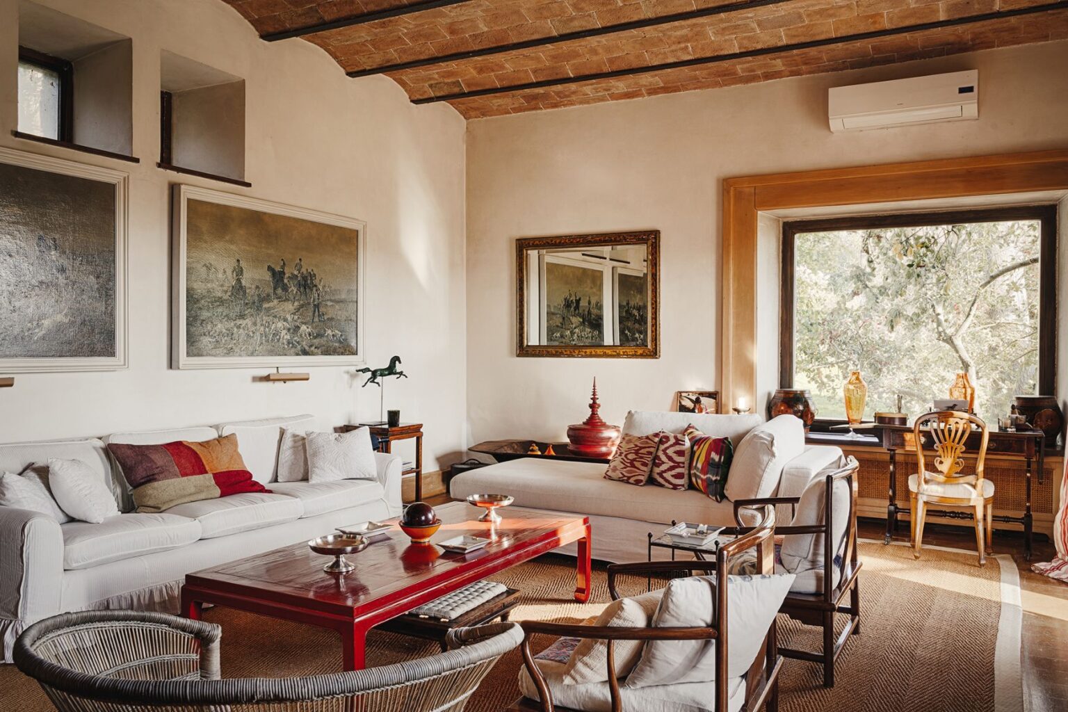Cozy living room featuring a mix of modern and vintage decor, including white sofas with colorful pillows, a red coffee table, and framed artwork on the walls. Natural light streams through a large window, illuminating the stylish furnishings and warm tones of the space. | Only on SopranoVillas