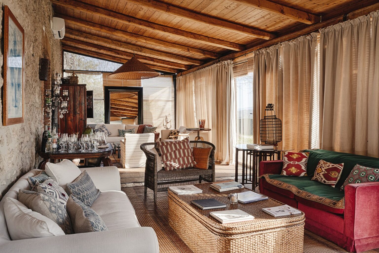 Cozy and stylish living room featuring a mix of modern and rustic decor, including a white sofa with patterned cushions, a wicker chair, and a red couch with decorative pillows. The room is illuminated by natural light through large windows, complemented by wooden beams and a woven ceiling pendant. A coffee table with books and glassware enhances the inviting atmosphere. | Only on SopranoVillas